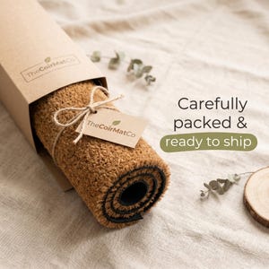 May include: A rolled-up brown coir doormat in a cardboard box, tied with twine and a tag. The tag and box feature the logo "TheCoirMatCo". The text "Carefully packed & ready to ship" is visible.