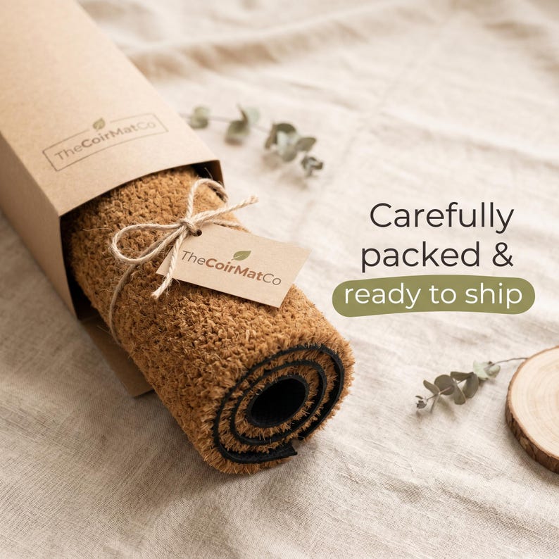 May include: A rolled-up brown coir doormat in a cardboard box, tied with twine and a tag. The tag and box feature the logo "TheCoirMatCo". The text "Carefully packed & ready to ship" is visible.