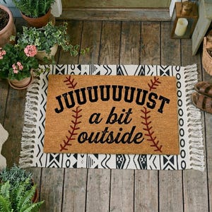 Puede incluir: Una alfombra rectangular con el texto "JUST a bit outside" en negro, diseñada para parecerse a una pelota de béisbol. La alfombra es marrón con detalles de costura rojos y está bordeada por una alfombra con estampado en blanco y negro con flecos.