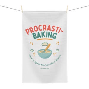 May include: White kitchen towel with the words "PROCRASTI-BAKING" in red and green. A cartoon illustration of a bowl with a whisk, clouds, and stars is below the text. The towel is hanging from a clothesline.