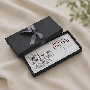 Könnte beinhalten: Eine schwarze rechteckige Geschenkbox mit grauem Band, die eine weiße Karte mit dem Text DINNER FOR TWO, HAPPY ANNIVERSARY, CLARA, THREE COURSES BY CANDLELIGHT, BATH, 14 MAY 2027, 7:30 PM, LOVE, JAMES X und einer Skizze eines Gedecks enthält.