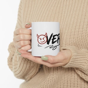 May include: White ceramic mug with the word "OVER" in black script, accompanied by a red devil face with horns. The mug is held by a person wearing a beige sweater.