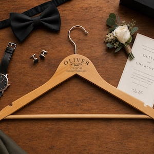 May include: A wooden clothes hanger engraved with "Oliver Groom 22nd September 2027" is surrounded by wedding accessories. A black bow tie, watch, cufflinks, and invitation card are arranged on a wooden surface, with a small floral arrangement.