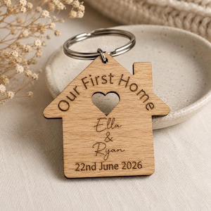 Könnte beinhalten: Ein holzfarbener Schlüsselanhänger in Hausform mit der Aufschrift "Our First Home" und einem herzförmigen Ausschnitt. Die Namen "Ella & Ryan" und das Datum "22nd June 2026" sind eingraviert. Befestigt an einem silbernen Metallring.