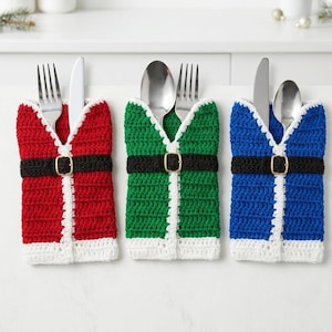 May include: Three crochet silverware holders in red, green, and blue. Each holder features a white trim, a black belt with a gold-colored buckle, and contains a metal fork, spoon, or knife. The holders are arranged side by side on a flat surface.