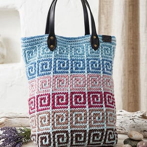 May include: A crochet tote bag featuring a gradient of square patterns in shades of blue, pink, magenta, and brown. The bag has two black leather handles attached with brass rivets and a small rectangular tag on the upper right side of the exterior.