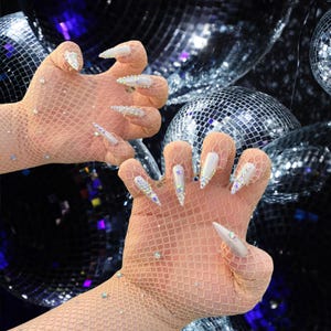 Peut inclure: Gros plan sur deux mains gantées de résille beige ornées de strass irisés. Les longs ongles pointus sont peints en blanc et ornés de strass. L'arrière-plan présente plusieurs boules à facettes.