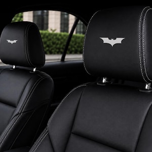 Pode incluir: Dois encostos de cabeça de assento de carro em couro preto com logotipos do Batman bordados em branco. Os encostos de cabeça têm costuras brancas visíveis ao longo das bordas e são montados em hastes de metal para o interior do veículo.