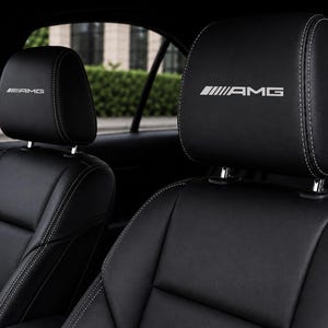 Puede incluir: Asientos de coche de cuero negro con costuras blancas en contraste y el logotipo AMG bordado en blanco en los reposacabezas. Los asientos tienen una textura suave y un diseño estructurado con costuras visibles a lo largo de los bordes.