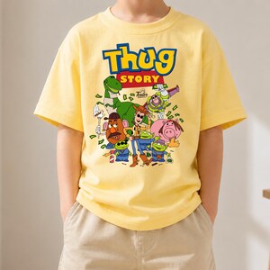 May include: A light yellow short-sleeved t-shirt featuring a graphic print with the text "Thug Story" in blue and yellow letters. The design includes various cartoon characters, including a green dinosaur, a cowboy, and aliens, surrounded by green money.