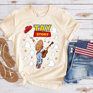 May include: A cream-colored short-sleeve t-shirt featuring a graphic of a person with long hair seen from behind, wearing cow-print pants and holding a guitar. The text TAY STORY is printed above, accompanied by a red cowboy hat, clouds, and stars.