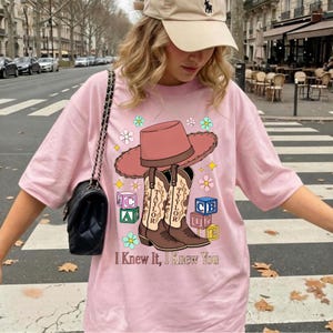 May include: A light pink t-shirt featuring a graphic of a pink cowboy hat, two brown cowboy boots labeled Taylor, colorful alphabet blocks, and flowers. The text "I Knew It, I Knew You" is printed at the bottom of the graphic design on the shirt.
