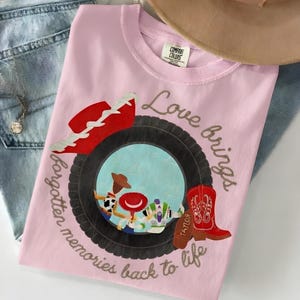 May include: A light pink t-shirt featuring a graphic of a black tire framing a scene with three cartoon characters. A red cowboy hat sits above the tire, and a red cowboy boot is to the right. The text reads, Love brings forgotten memories back to life.