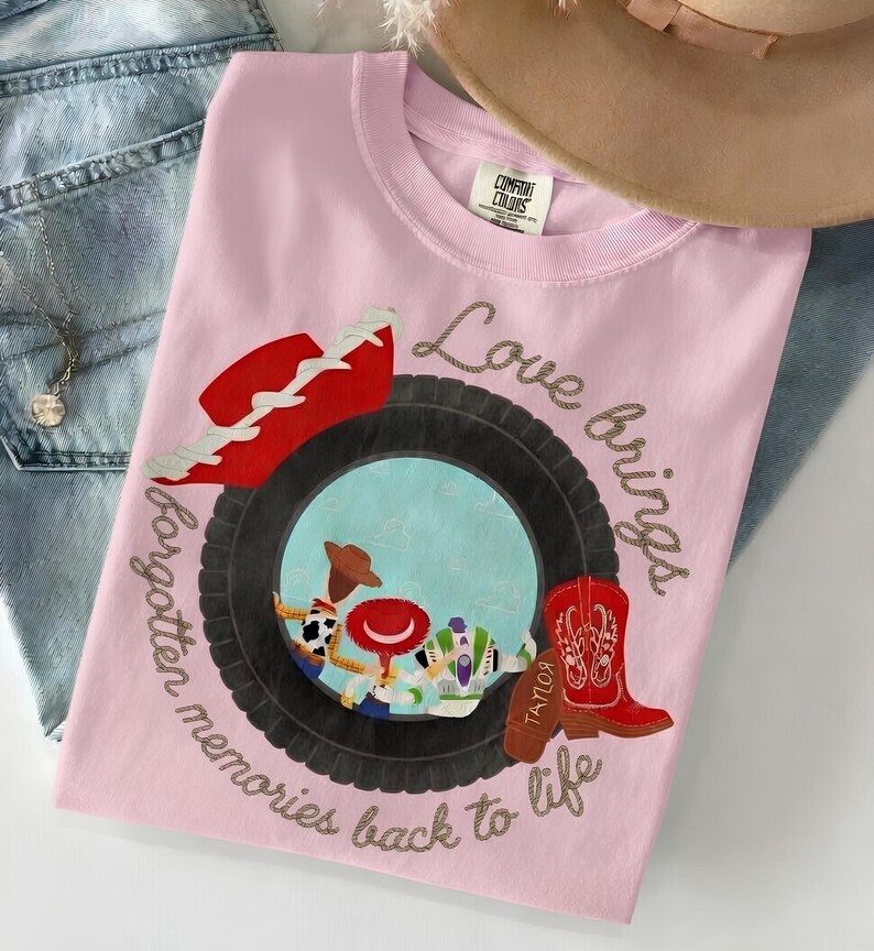 May include: A light pink t-shirt featuring a graphic of a black tire framing a scene with three cartoon characters. A red cowboy hat sits above the tire, and a red cowboy boot is to the right. The text reads, Love brings forgotten memories back to life.