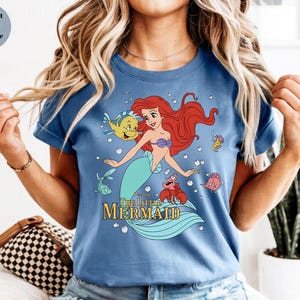 Może przedstawiać: Błękitny t-shirt z nadrukiem graficznym przedstawiającym Arielkę z Małej Syrenki wraz z jej przyjaci&oacute;łmi Florkiem i Sebastianem. Wz&oacute;r zawiera bąbelki oraz napis The Little Mermaid ż&oacute;łtymi literami. Koszulka ma kr&oacute;tkie rękawy i okrągły dekolt.