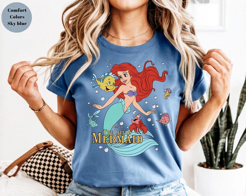 Może przedstawiać: Błękitny t-shirt z nadrukiem graficznym przedstawiającym Arielkę z Małej Syrenki wraz z jej przyjaci&oacute;łmi Florkiem i Sebastianem. Wz&oacute;r zawiera bąbelki oraz napis The Little Mermaid ż&oacute;łtymi literami. Koszulka ma kr&oacute;tkie rękawy i okrągły dekolt.