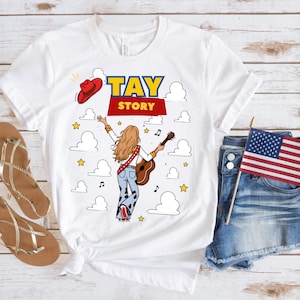 May include: A white t-shirt featuring a graphic of a person with long blonde hair seen from behind, wearing light blue cow-print bell-bottom jeans and holding an acoustic guitar. The text TAY STORY is printed above, with a red cowboy hat, clouds, and stars.