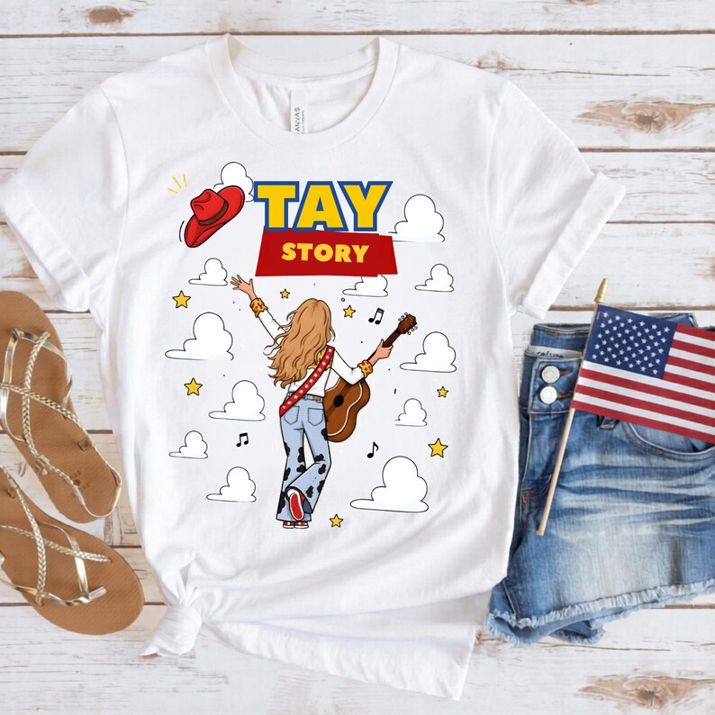 May include: A white t-shirt featuring a graphic of a person with long blonde hair seen from behind, wearing light blue cow-print bell-bottom jeans and holding an acoustic guitar. The text TAY STORY is printed above, with a red cowboy hat, clouds, and stars.