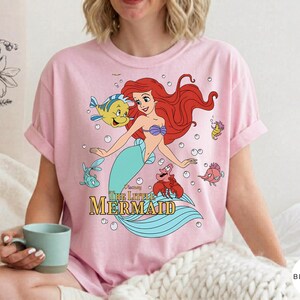 Może przedstawiać: Jasnor&oacute;żowy t-shirt z kr&oacute;tkim rękawem z nadrukiem graficznym przedstawiającym syrenkę Ariel wraz z Florkiem i Sebastianem. Poniżej postaci znajduje się napis "Disney The Little Mermaid" wykonany ż&oacute;łtymi i złotymi literami.