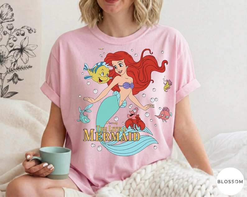 Może przedstawiać: Jasnor&oacute;żowy t-shirt z kr&oacute;tkim rękawem z nadrukiem graficznym przedstawiającym syrenkę Ariel wraz z Florkiem i Sebastianem. Poniżej postaci znajduje się napis "Disney The Little Mermaid" wykonany ż&oacute;łtymi i złotymi literami.