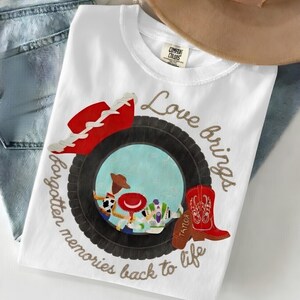 May include: A white t-shirt featuring a graphic of a black tire surrounding a blue sky scene with cartoon characters. A red cowboy hat and a red cowboy boot are positioned on the tire. The text reads Love brings forgotten memories back to life.