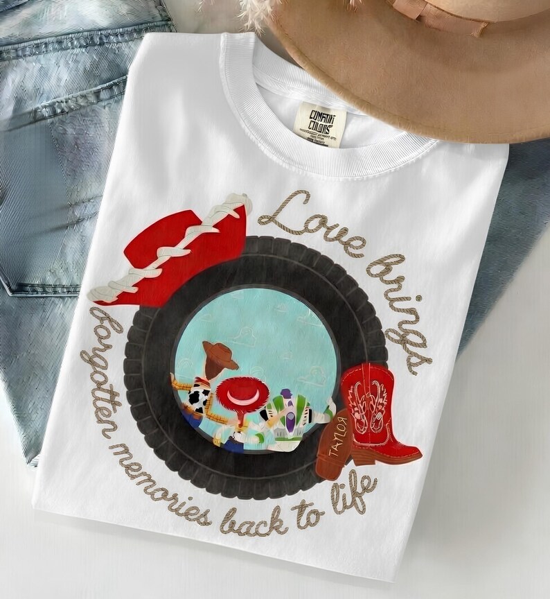 May include: A white t-shirt featuring a graphic of a black tire surrounding a blue sky scene with cartoon characters. A red cowboy hat and a red cowboy boot are positioned on the tire. The text reads Love brings forgotten memories back to life.