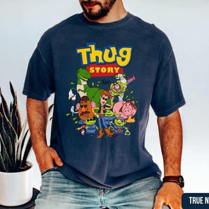 May include: A navy blue short-sleeve t-shirt featuring a graphic print with the text "THUG STORY" in yellow and red. The design includes cartoon characters such as a dinosaur, a space ranger, a cowboy, a potato figure, a pig, and green aliens.