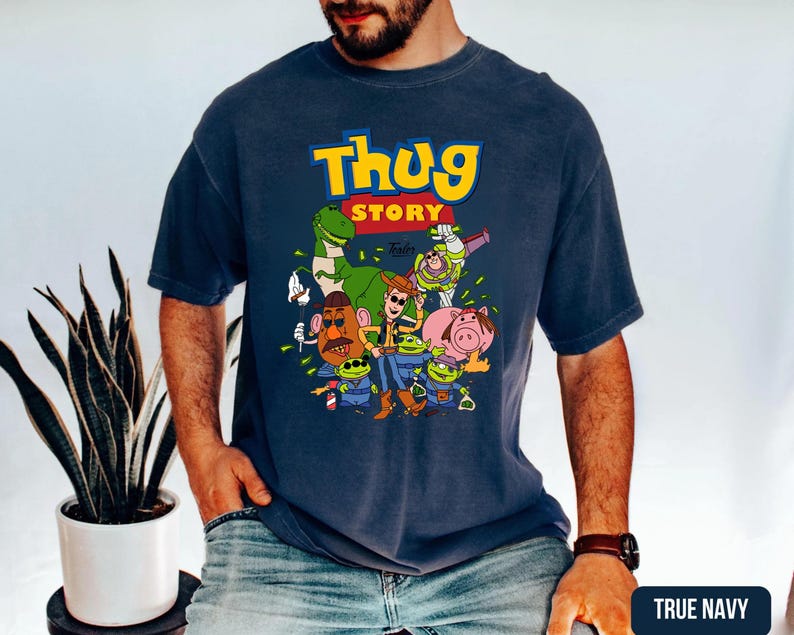 May include: A navy blue short-sleeve t-shirt featuring a graphic print with the text "THUG STORY" in yellow and red. The design includes cartoon characters such as a dinosaur, a space ranger, a cowboy, a potato figure, a pig, and green aliens.