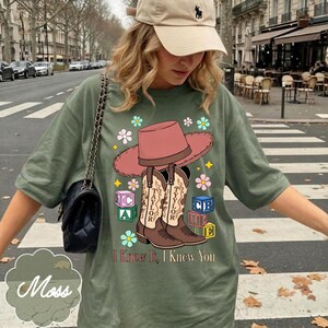 May include: An olive green t-shirt featuring a graphic print of a pink cowboy hat, two brown cowboy boots labeled Taylor, colorful alphabet blocks, and flowers. The text I Knew It, I Knew You is printed in brown below the boots and blocks.