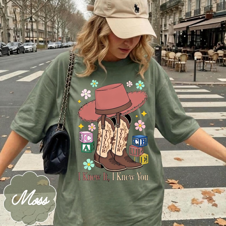 May include: An olive green t-shirt featuring a graphic print of a pink cowboy hat, two brown cowboy boots labeled Taylor, colorful alphabet blocks, and flowers. The text I Knew It, I Knew You is printed in brown below the boots and blocks.
