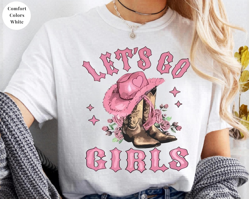 May include: A white t-shirt featuring a graphic print with the text "LET'S GO GIRLS" in pink letters. The design includes a pink cowboy hat with a heart, brown cowboy boots, and pink floral accents, all arranged in a central composition.