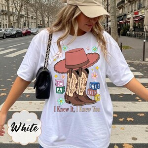 May include: A white t-shirt featuring a graphic print of a pink cowboy hat resting on two brown cowboy boots, surrounded by colorful alphabet blocks and flowers. The text "I Knew It, I Knew You" is printed below the boots in a brown serif font.