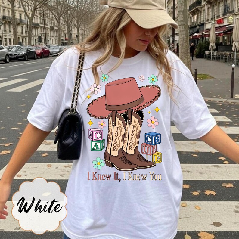 May include: A white t-shirt featuring a graphic print of a pink cowboy hat resting on two brown cowboy boots, surrounded by colorful alphabet blocks and flowers. The text "I Knew It, I Knew You" is printed below the boots in a brown serif font.