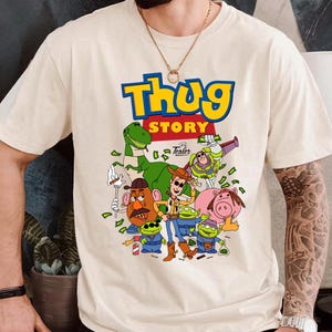 May include: A cream-colored short-sleeve t-shirt featuring a graphic print with the text "Thug Story" in blue and yellow. The design includes cartoon characters such as a cowboy, a dinosaur, a space ranger, a potato figure, and three aliens.