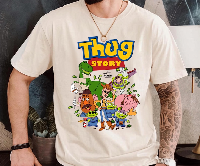 May include: A cream-colored short-sleeve t-shirt featuring a graphic print with the text "Thug Story" in blue and yellow. The design includes cartoon characters such as a cowboy, a dinosaur, a space ranger, a potato figure, and three aliens.
