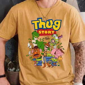 May include: A mustard yellow short-sleeve t-shirt featuring a graphic print with the text "Thug Story" in blue and yellow letters. The illustration depicts various cartoon characters including a dinosaur, a cowboy, and aliens with money.