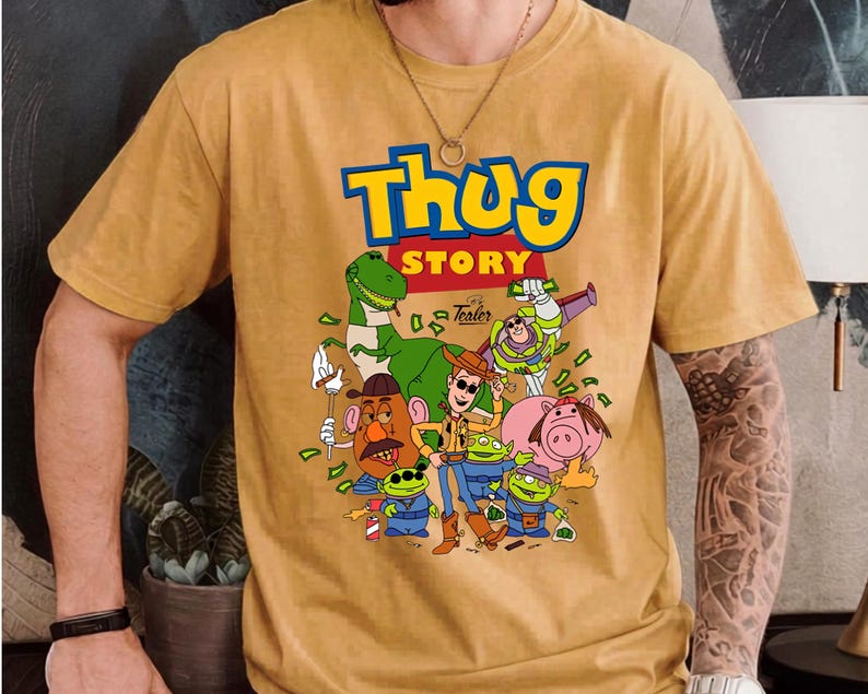 May include: A mustard yellow short-sleeve t-shirt featuring a graphic print with the text "Thug Story" in blue and yellow letters. The illustration depicts various cartoon characters including a dinosaur, a cowboy, and aliens with money.