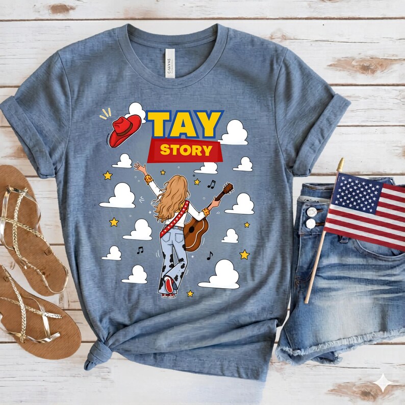 May include: A heather blue t-shirt featuring a graphic of a person with long blonde hair playing an acoustic guitar, wearing cow-print pants and a red hat. The text TAY STORY is printed above, surrounded by white clouds, yellow stars, and musical notes.