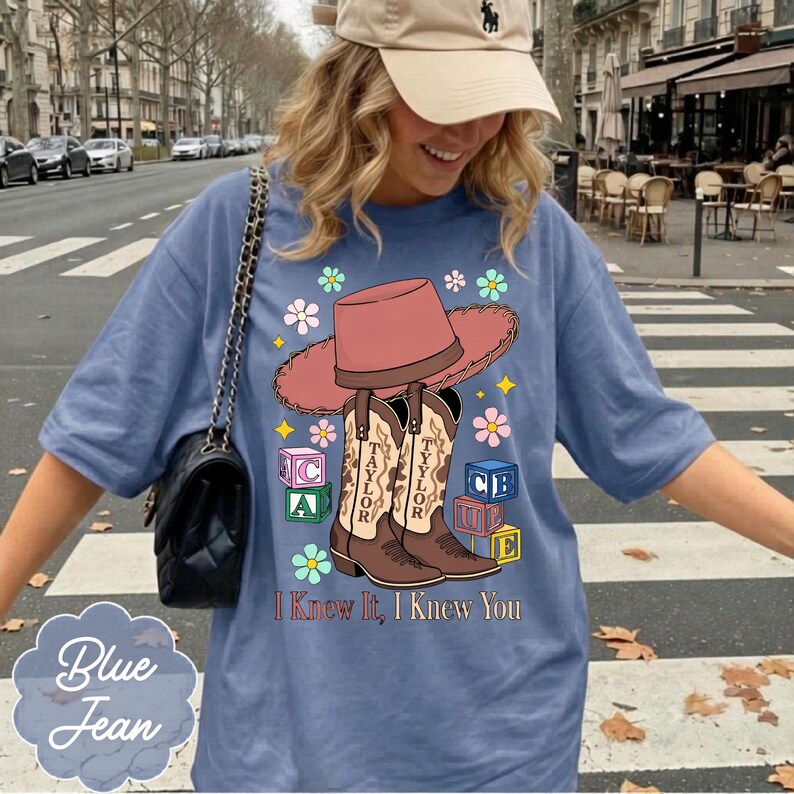 May include: A blue t-shirt featuring a graphic of a pink cowboy hat, two brown cowboy boots labeled Taylor, colorful alphabet blocks, and small flowers. The text I Knew It, I Knew You is printed below the boots in a simple, dark font.
