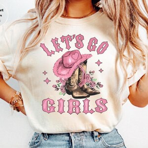 May include: An ivory t-shirt featuring a graphic print with the text "LET'S GO GIRLS" in pink. The design includes a pink cowboy hat with a heart icon, brown cowboy boots, and pink floral accents, surrounded by small four-pointed star shapes.