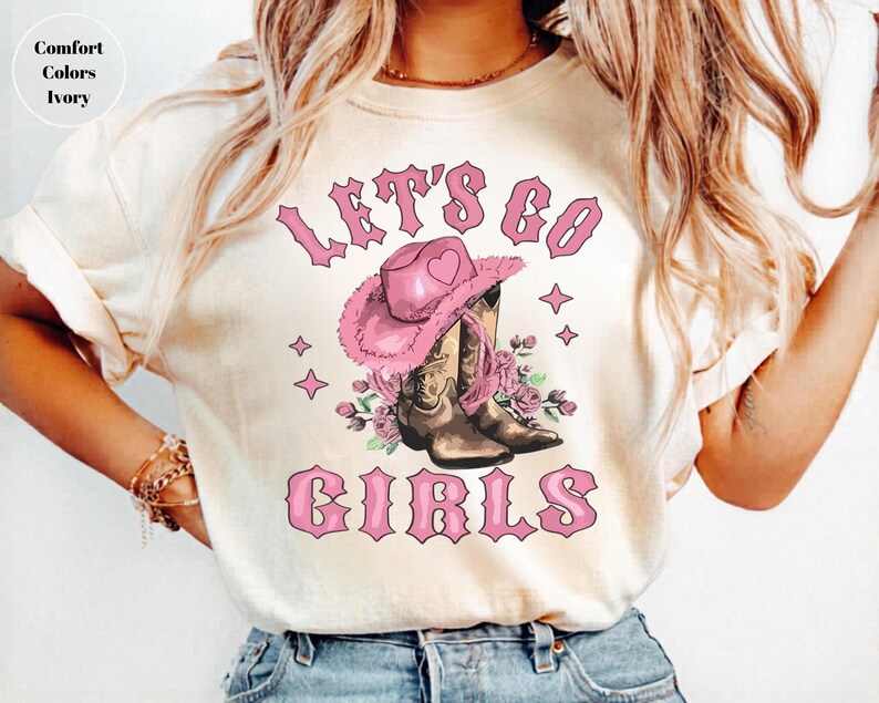 May include: An ivory t-shirt featuring a graphic print with the text "LET'S GO GIRLS" in pink. The design includes a pink cowboy hat with a heart icon, brown cowboy boots, and pink floral accents, surrounded by small four-pointed star shapes.