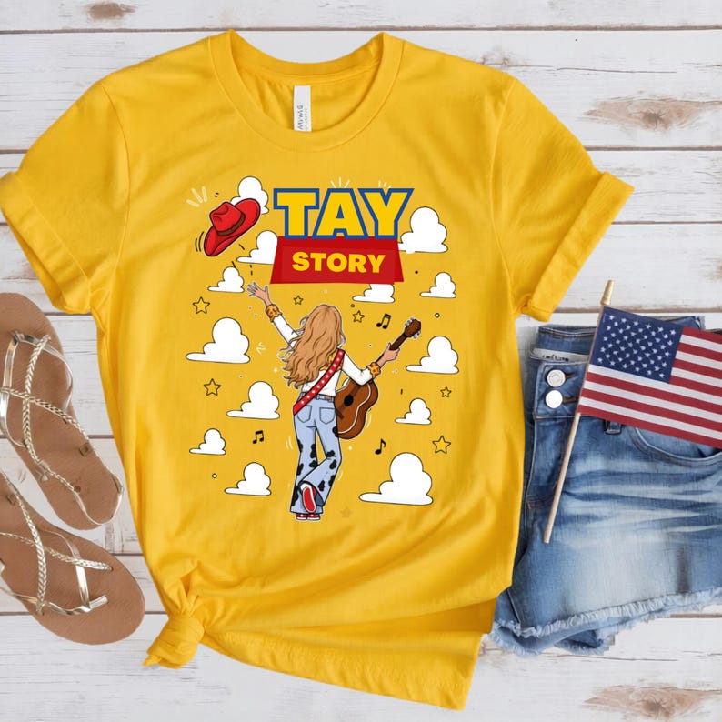 May include: A bright yellow short-sleeve t-shirt featuring a graphic print of a person with long hair playing an acoustic guitar, wearing cow-print pants and a red cowboy hat. The text TAY STORY is printed above, surrounded by white clouds and stars.