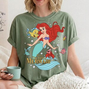 Może przedstawiać: Oliwkowozielony t-shirt z kr&oacute;tkim rękawem z nadrukiem graficznym przedstawiającym Arielkę z Małej Syrenki, wraz z Florkiem, Sebastianem i innymi stworzeniami morskimi. Napis "Disney The Little Mermaid" jest wydrukowany złotymi literami pod postaciami.