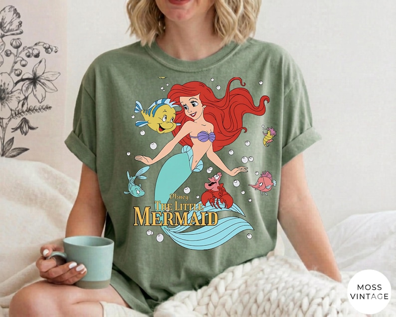 Może przedstawiać: Oliwkowozielony t-shirt z kr&oacute;tkim rękawem z nadrukiem graficznym przedstawiającym Arielkę z Małej Syrenki, wraz z Florkiem, Sebastianem i innymi stworzeniami morskimi. Napis "Disney The Little Mermaid" jest wydrukowany złotymi literami pod postaciami.