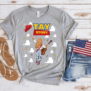 May include: A heather gray t-shirt featuring a graphic of a person with long blonde hair seen from behind, playing an acoustic guitar. The design includes the text TAY STORY, a red cowboy hat, white clouds, yellow stars, and musical notes.