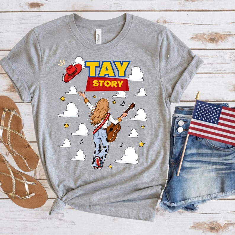 May include: A heather gray t-shirt featuring a graphic of a person with long blonde hair seen from behind, playing an acoustic guitar. The design includes the text TAY STORY, a red cowboy hat, white clouds, yellow stars, and musical notes.