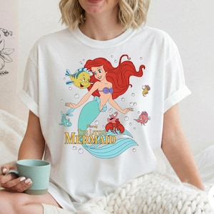Może przedstawiać: Biały t-shirt z kolorową grafiką przedstawiającą Ariel z Małej Syrenki wraz z Florkiem, Sebastianem i innymi morskimi stworzeniami. Wz&oacute;r zawiera bąbelki oraz napis Disney The Little Mermaid ż&oacute;łtymi i czarnymi literami.