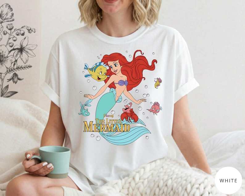Może przedstawiać: Biały t-shirt z kolorową grafiką przedstawiającą Ariel z Małej Syrenki wraz z Florkiem, Sebastianem i innymi morskimi stworzeniami. Wz&oacute;r zawiera bąbelki oraz napis Disney The Little Mermaid ż&oacute;łtymi i czarnymi literami.