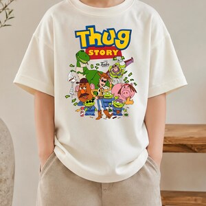 May include: An off-white short-sleeve t-shirt featuring a graphic print with the text "Thug Story" in blue and yellow. The design includes cartoon characters like a cowboy, a space ranger, a dinosaur, a potato figure, and green aliens.