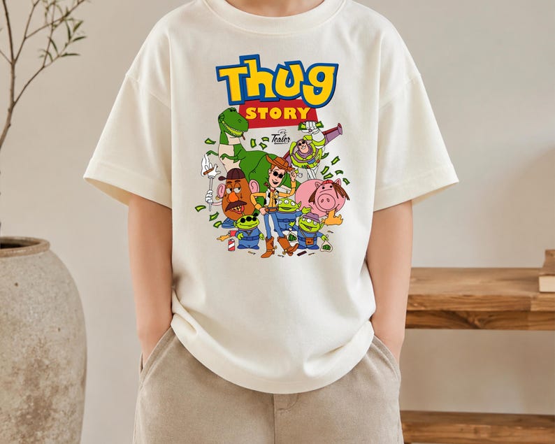 May include: An off-white short-sleeve t-shirt featuring a graphic print with the text "Thug Story" in blue and yellow. The design includes cartoon characters like a cowboy, a space ranger, a dinosaur, a potato figure, and green aliens.